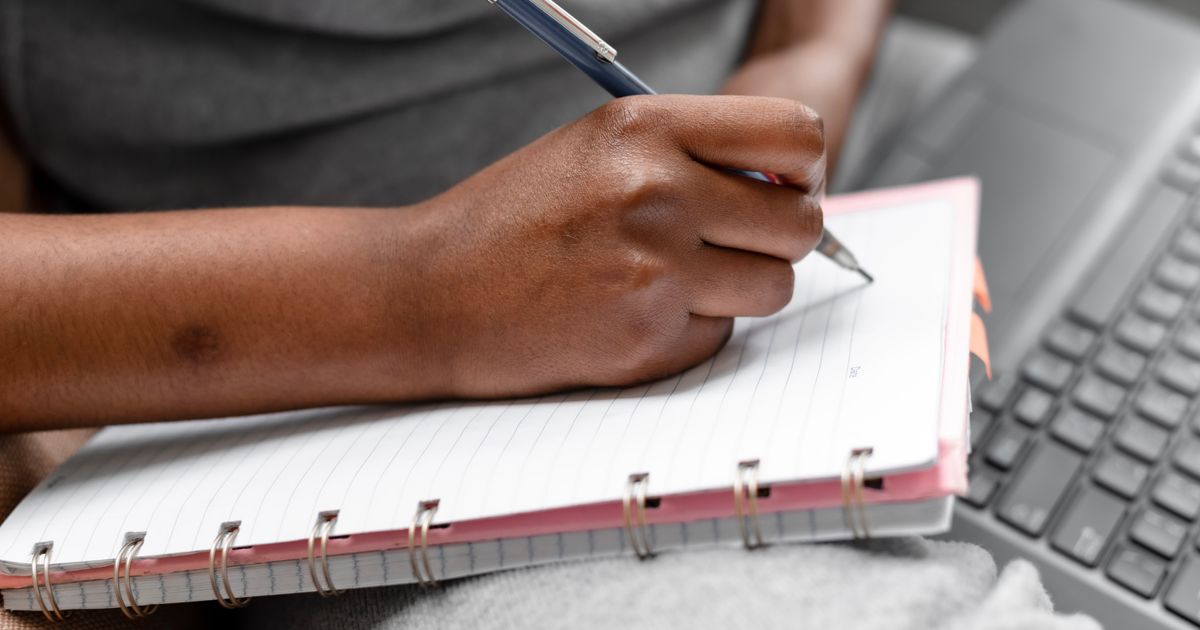 A person making notes in a spiral notebook beside a laptop